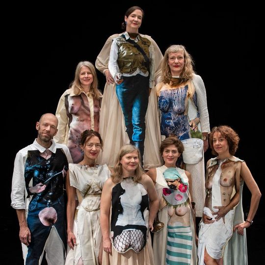 Auf dem Foto stehen die 8 Performer_innen von She She Pop in einer Pyramiden-Konstellation. 5 stehen unten in der ersten Reihe, zwei in der oberen zweiten Reihe und zwei ganz oben in der Spitze der menschlichen Pyramide. Alle Performer_innen tragen bunte Kostüme und schauen den Bild-Betrachtenden lächelnd an.