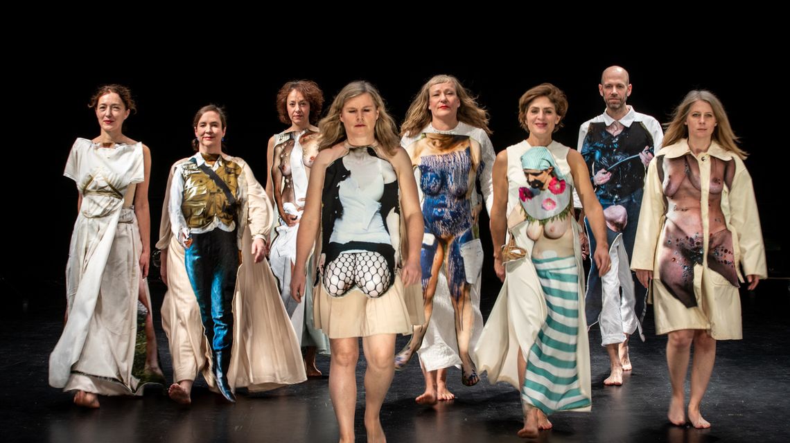 Auf dem Inszenierungsfoto einer Show von SHE SHE POP sind 8 Performer_innen der Gruppe zu sehen, die auf den Betrachtenden zulaufen. Die Performer_innen haben bunte Bühnenkostüme an, manche lächeln, manche schauen dem Betrachtenden ernst entgegen.