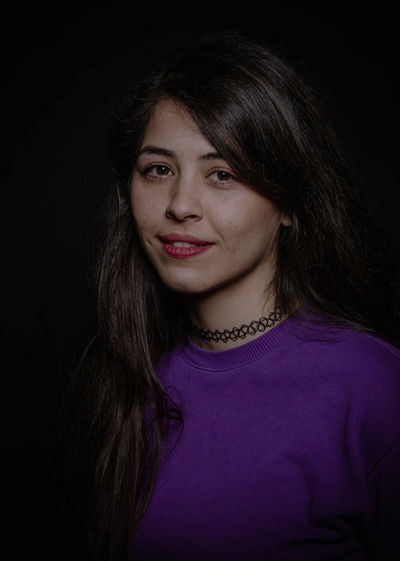 Das Porträt zeigt die Dozentin Betül Çınar. Sie hat langes, schwarzes Haar und trägt einen lilafarbenen Pullover und ein schwarzes Tattoo-Halsband. Sie lächelt.